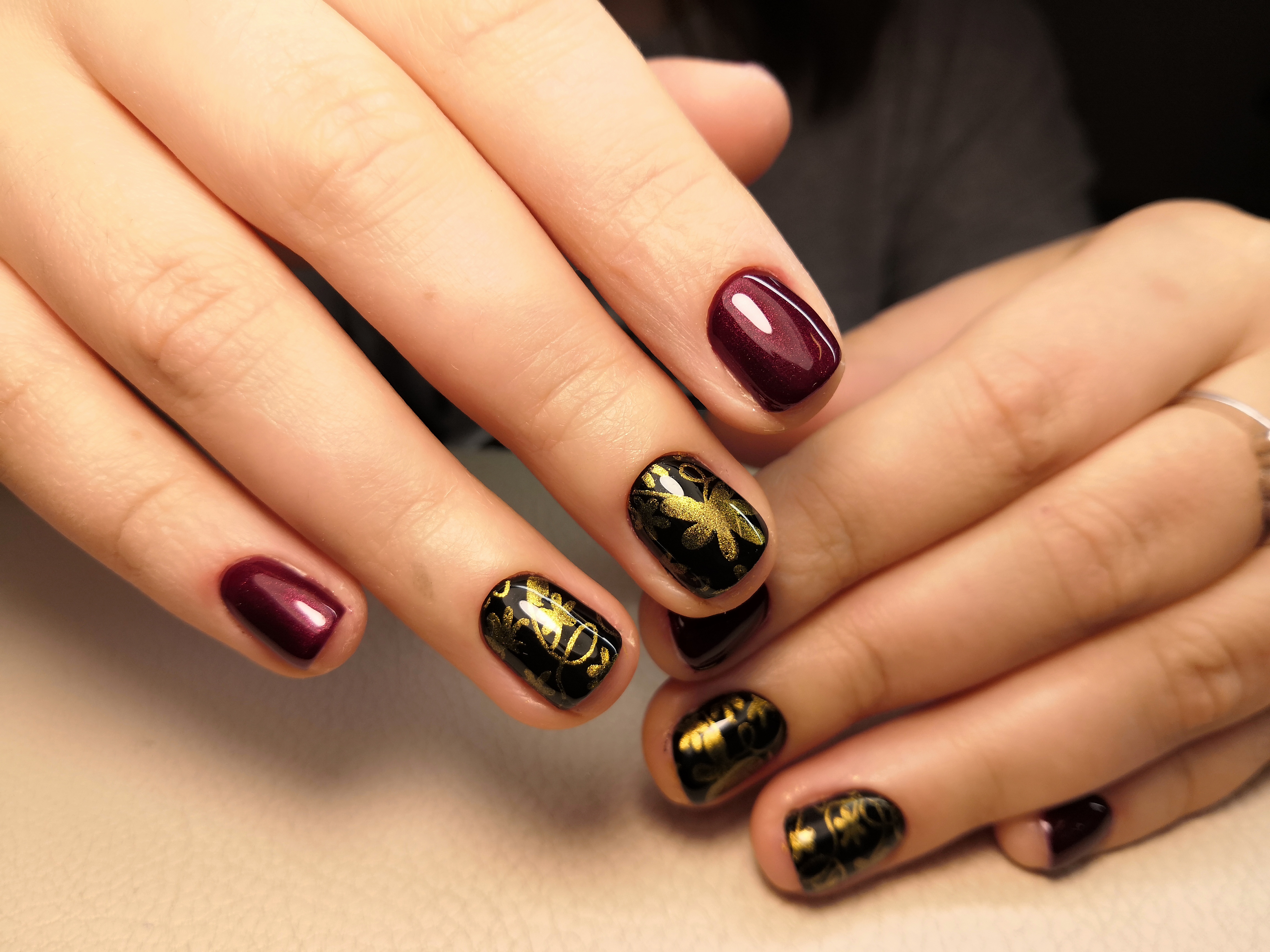 Маникюр в тёмно-баклажановом цвете с чёрным дизайном и золотыми блестящими рисунками.